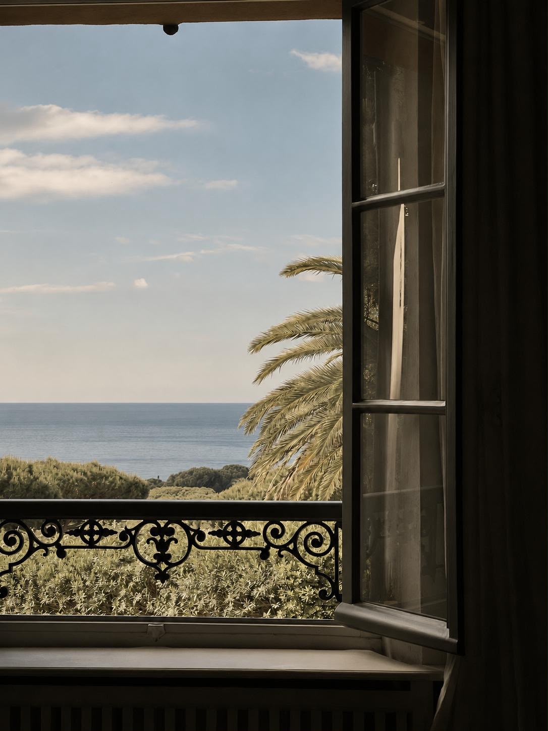 Fenêtre à cadre sombre et garde-corps en fer forgé ouverte sur un paysage de mer et de palmier, illustrant la relation entre intérieur et extérieur